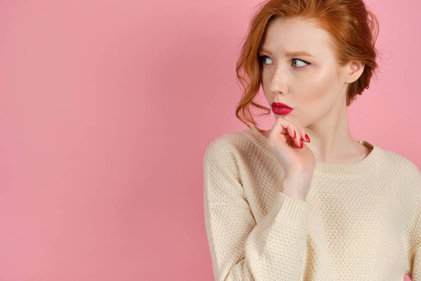 A red-haired girl in a light sweater with red lips stands on a pink background and looks warily to the side, propping her chin. - Foto, immagini