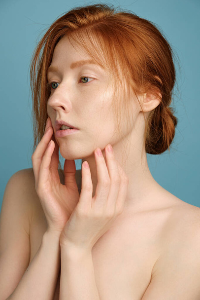  Redhead girl with clean radiant skin, no makeup, standing in a half-turn on a blue background, putting his palms to his face - Foto, Bild