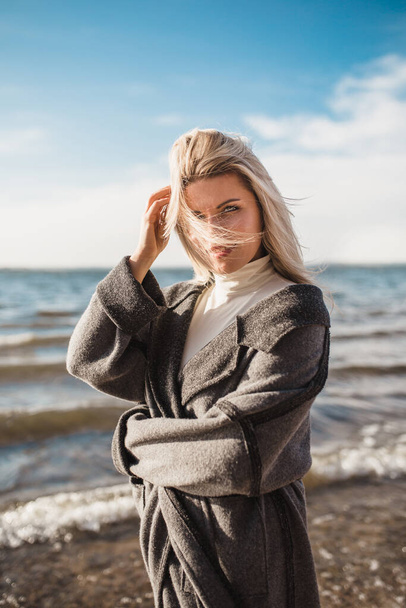 A delightful happy sensual seductive Scandinavian blonde with beautiful eyes and a white-toothed smile wraps herself comfortably in a wool coat on the shore of a harsh sea - Foto, afbeelding