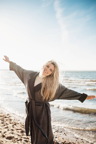 Delightful happy slender Scandinavian blonde with beautiful eyes and a white-toothed smile merrily spread her arms to the sides on the seashore - Фото, зображення