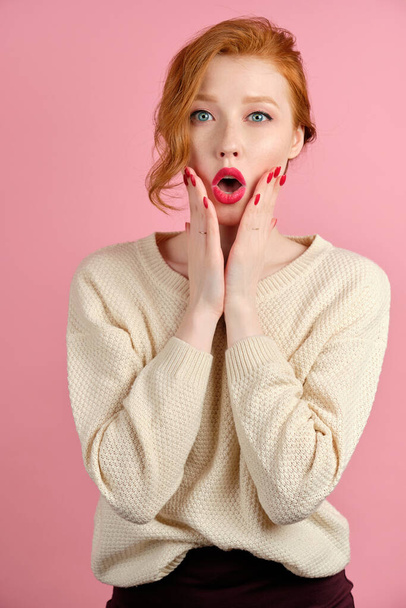 A red-haired girl with red lips, stands on a pink background, opening mouth in surprise and pressing hands to face - Zdjęcie, obraz
