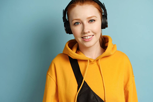 Red-haired girl in a yellow sweatshirt and headphones stands on a blue background and smiles - Valokuva, kuva