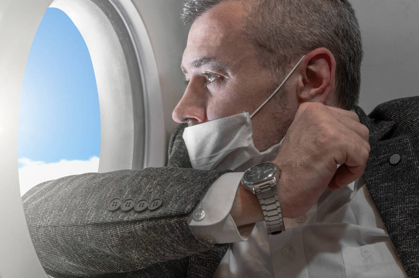 Ένας άντρας κοιτάζει μέσα στο φινιστρίνι ενός αεροπλάνου. Επιχειρηματίας με σακάκι και μάσκα από τον ιό.. - Φωτογραφία, εικόνα