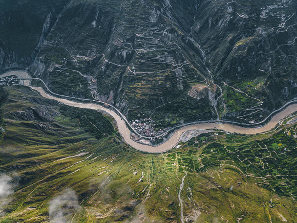 Top down vista aerea drone del fiume e piccolo insediamento tra le alte montagne nella zona tibetana della provincia del Sichuan, Cina. Scatto cinematografico, strade a serpentina curve e colline. Viaggio in primavera. - Foto, immagini
