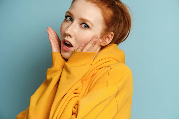 A red-haired girl in a yellow sweatshirt stands on a blue background and looks in surprise at the camera, raising palms to face - Фото, изображение