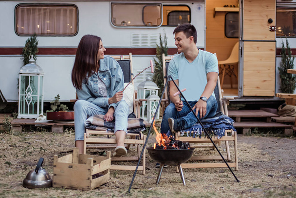 Happy young family at a campsite fries sausages on a bonfire. Picnic of a beautiful couple in love at the trailer, sitting comfortably, hugging, battle on sausages, kissing, laughing. Rest in nature - Photo, image