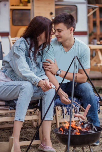 Happy young family at a campsite fries sausages on a bonfire. Picnic of a beautiful couple in love at the trailer, sitting comfortably, hugging, battle on sausages, kissing, laughing. Rest in nature - 写真・画像