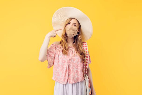 Happy young woman in a summer hat, with a protective medical mask on her face, on an yellow background - Foto, afbeelding