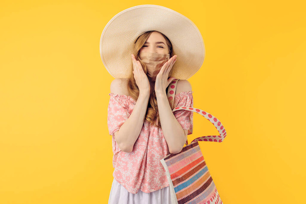 Happy excited young woman in a summer hat, with a protective medical mask on her face, on an isolated yellow background - Foto, immagini