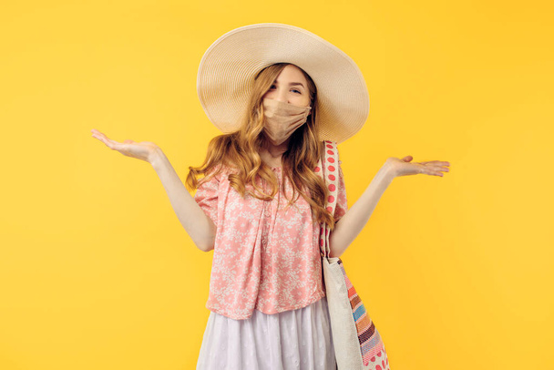 Happy excited young woman in a summer hat, with a protective medical mask on her face, on an isolated yellow background - Фото, изображение