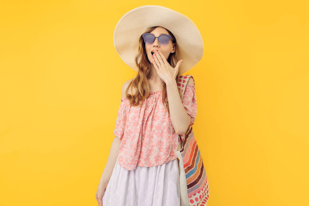 Uma jovem mulher atraente chocada e atenciosa em um chapéu de verão e óculos de sol, em um fundo amarelo isolado
 - Foto, Imagem