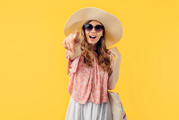 A happy excited young woman in a summer hat and sunglasses points at the camera on a yellow background - Foto, Imagem