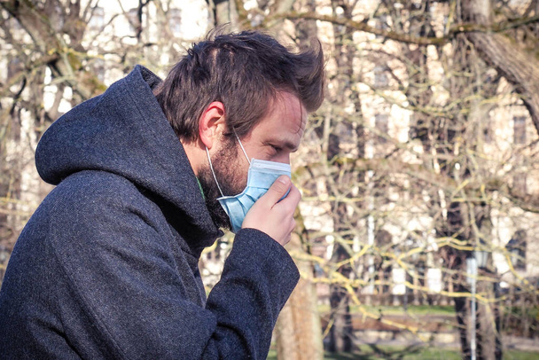 Handsome young European man coughing on the street with a medical face mask on. Closeup of a 35-year-old male in a respirator to protect against infection with influenza or coronavirus (Covid-19) - Photo, Image