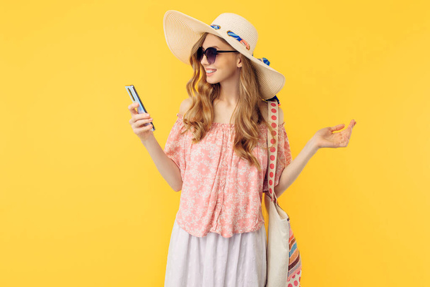 A happy smiling young woman in a summer hat and sunglasses, holding a smartphone on an isolated yellow background - Foto, Bild