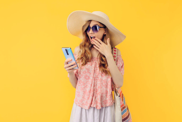 A shocked surprised young woman in a summer hat and sunglasses with a mobile phone on an isolated yellow background - Фото, зображення