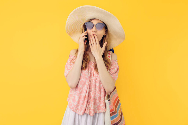 A shocked surprised young woman in a summer hat and sunglasses is talking on a mobile phone against an isolated yellow background - Photo, image