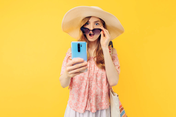 Yllättynyt nuori nainen kesähatussa ja aurinkolasit matkapuhelimella eristetyllä keltaisella taustalla - Valokuva, kuva