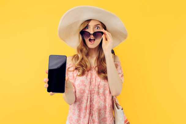 An excited shocked young woman in a summer hat and sunglasses shows a blank mobile phone screen for a copy of the space on an isolated yellow background - Foto, Bild