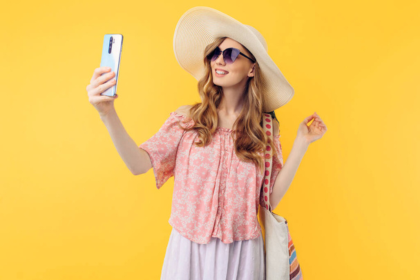 A happy smiling young woman in a summer hat and sunglasses takes a selfie on a mobile phone on an isolated pink background - Фото, изображение