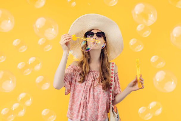 A happy attractive young woman in a summer hat blows soap bubbles on an isolated yellow background - Valokuva, kuva