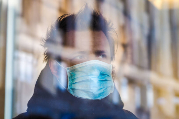 Красивый европейский мужчина стоит за стеклом в карантине в медицинской маске на лице. Крупный план 35-летнего мужчины в респираторе для защиты от инфекции коронавирусом (Covid-19). Дефокусировка
 - Фото, изображение
