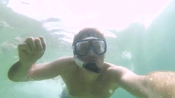 Gyönyörű fiatal srác búvármaszkban búvárkodik és integet a kamerának, miközben tiszta kék vízben úszik a tengeren. Víz alatti világ halakkal és korallzátonnyal. Az ember szelfit készít. - Felvétel, videó