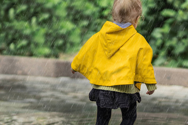 A joyful little girl in a yellow raincoat walking in the rain on the street alone. Park, nature, outdoors. Childhood concept. Universal Children's Day. - 写真・画像