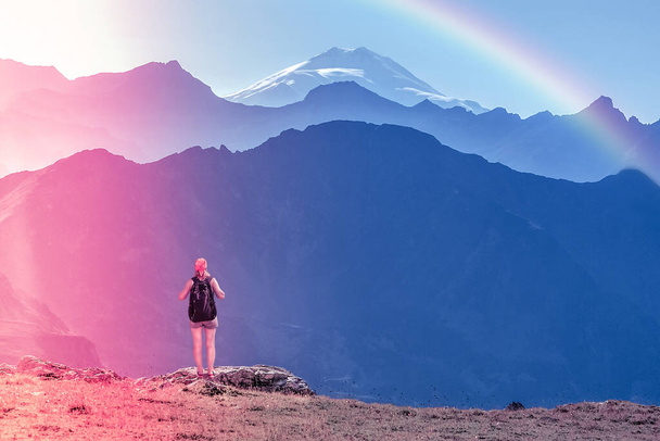 Girl traveler with backpack stands on a stone back to the viewer. She looks at the snow-capped top. Picturesque cloudless landscape with a rainbow. Wallpaper for concept of business thinking. climbing - Zdjęcie, obraz