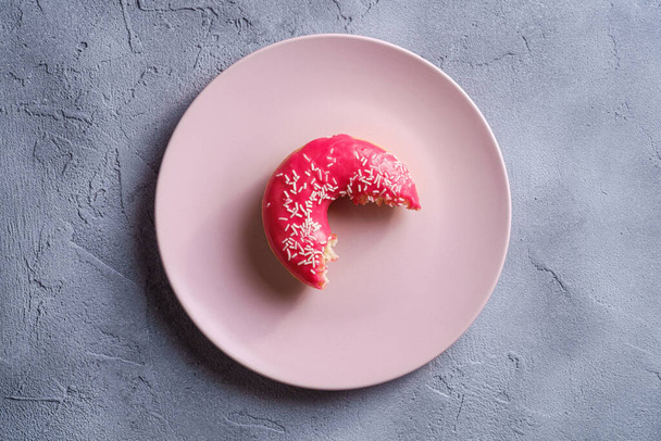 Кусатый розовый пончик с брызгами на розовую тарелку, сладкий застекленный десерт на бетонном текстурированном фоне, вид сверху
 - Фото, изображение