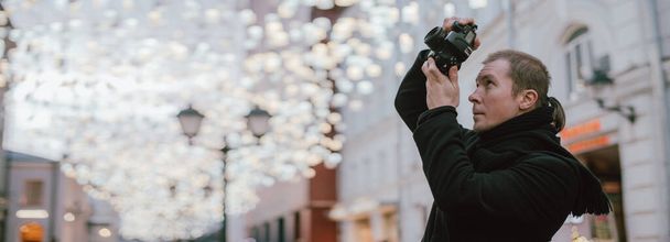 Un uomo con una macchina fotografica fotografa la strada della città vecchia in inverno. Fotografo con una macchina fotografica in cappotto e sciarpa sullo sfondo di lampioni decorati ad arte - Foto, immagini
