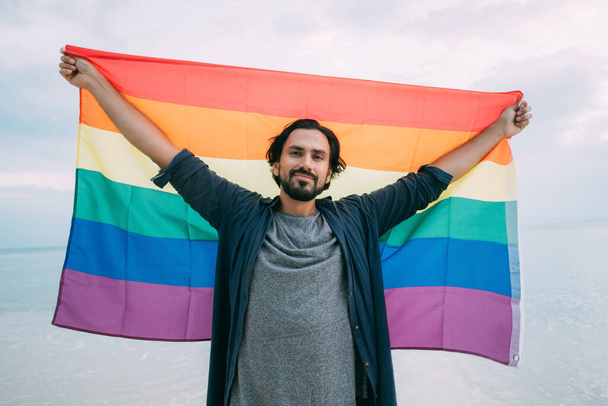 Un tipo alegre con una bandera de arco iris en la playa. Joven sosteniendo una bandera del arco iris contra el cielo del océano
 - Foto, Imagen