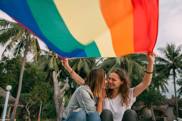 Duas mulheres com bandeira do arco-íris na praia em um fundo de palmeiras. Casal Meninas lésbicas jovens segurando uma bandeira do arco-íris e abraçando alegremente em uma ilha tropical
 - Foto, Imagem