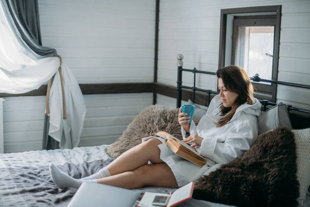 Una mujer está acostada en una cama con un libro. Niña descansando, leyendo un libro en el dormitorio en una gran cama acogedora por la tarde - Foto, Imagen