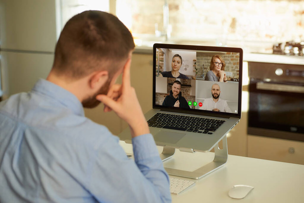 Πίσω όψη ενός αρσενικού υπαλλήλου που δουλεύει εξ αποστάσεως ακούγοντας τους συναδέλφους του για δουλειές σε μια τηλεδιάσκεψη σε έναν φορητό υπολογιστή στο σπίτι. Μια πολυεθνική επιχειρηματική ομάδα σε μια online συνάντηση. - Φωτογραφία, εικόνα