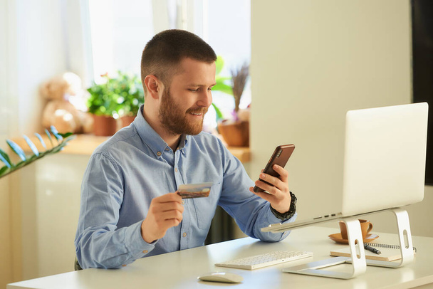 Ένας άνθρωπος που ψάχνει για προϊόντα για να αγοράσει σε ένα ηλεκτρονικό κατάστημα στο smartphone του με μια πιστωτική κάρτα στο σπίτι. Ένας τύπος με γενειάδα κάνει μια ηλεκτρονική πληρωμή στο διαδίκτυο σε ένα φορητό υπολογιστή στο διαμέρισμά του. - Φωτογραφία, εικόνα