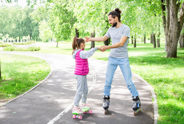 Perheaktiviteetteja kesäpuistossa. Onnellinen isä opettaa pientä tytärtä rullaluistelemaan puistossa. Isän päivän käsite. - Valokuva, kuva
