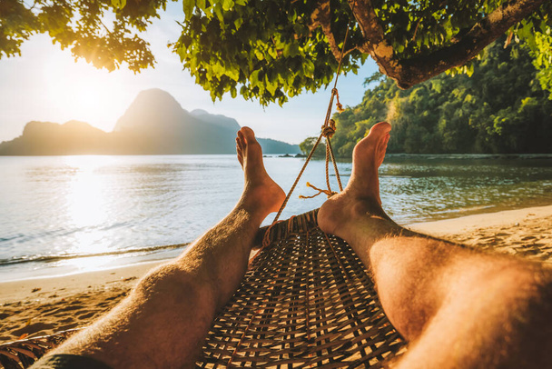 Feet of adult man relaxing in a hammock on the beach during summer holiday - Фото, изображение