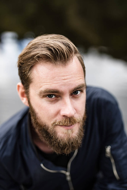 A really attractive Scandinavian dude with great facial features, hairand unshaven beard, wearing a blue jacket, spending his time bythe lake staring at the camera. - Foto, Bild