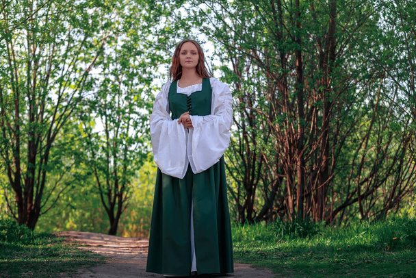 Krásná středověká mladá žena v tradičním historickém ženském zeleném a bílém kostýmu v přírodě. Fantazie dívka v dlouhých šatech nebo šaty chůze na cestě mezi stromy v lese. - Fotografie, Obrázek