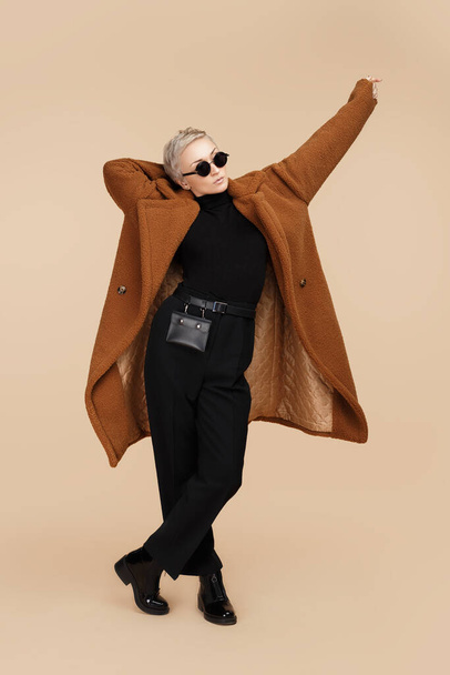 Confident young hipster woman with blonde short hair wearing a coat and sunglasses walking over beige background. - Foto, Imagen