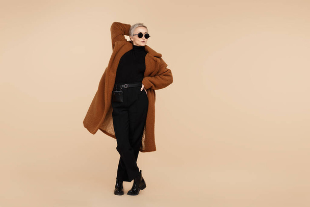 Trendy young hipster woman with blonde short hair wearing a coat and sunglasses walking over beige background. - Zdjęcie, obraz