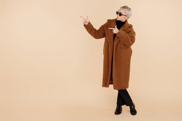 Hipster woman with blonde short hair wearing a coat and sunglasses point finger away isolated over beige background. - Foto, Imagen