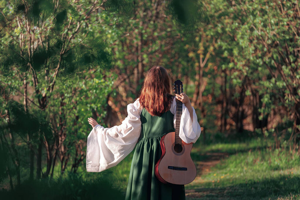 Młoda kobieta w średniowiecznej sukience spaceruje z gitarą w słonecznym lesie. Dziewczyna Fantasy lub bard w letniej przyrodzie. Widok z tyłu. - Zdjęcie, obraz