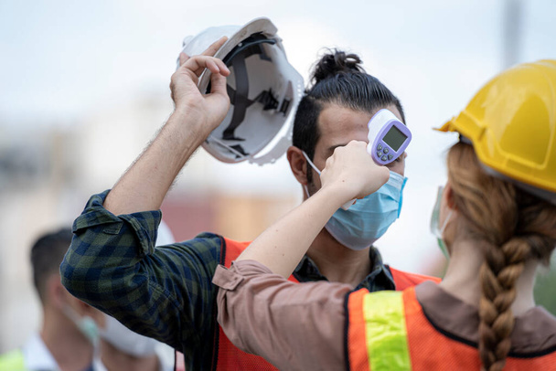 Factory woman worker in a face medical mask and safety dress used measures temperature at worker people standing on queue with a non-contact infrared thermometer - Photo, Image