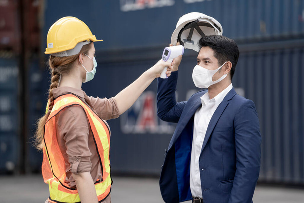Eine Fabrikarbeiterin in medizinischer Gesichtsmaske und Schutzkleidung misst mit einem berührungslosen Infrarot-Thermometer die Temperatur der Arbeiter, die in der Schlange stehen. Hintergrund des Frachtcontainers - Foto, Bild