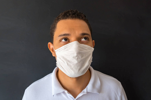 Portrait of a handsome young man with a surgical medical mask on a white shirt, standing up. Indoor studio shot, isolated on a black background. - Photo, image
