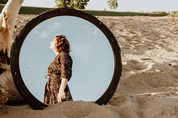 Outdoor atmospheric lifestyle photo of young beautiful  darkhaired woman in dress  and jacket posing on the beach in the reflection of the mirror.Conceptual photo with a mirror. - Foto, Bild