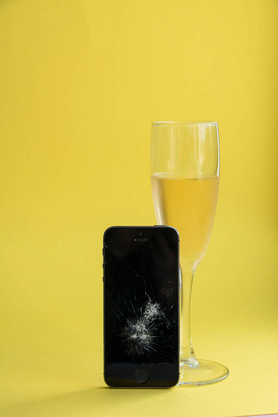 Разбитый черный смартфон с бокалом шампанского на ярко-желтом фоне
 - Фото, изображение