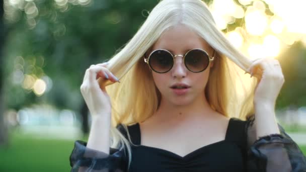 Roztomilé mladé kavkazské blondýny dívka v kulaté stylové sluneční brýle a černé šaty účinně nadýmá její dlouhé vlasy s rukama s manikúrou na pozadí stromů a trávy městského parku. - Záběry, video