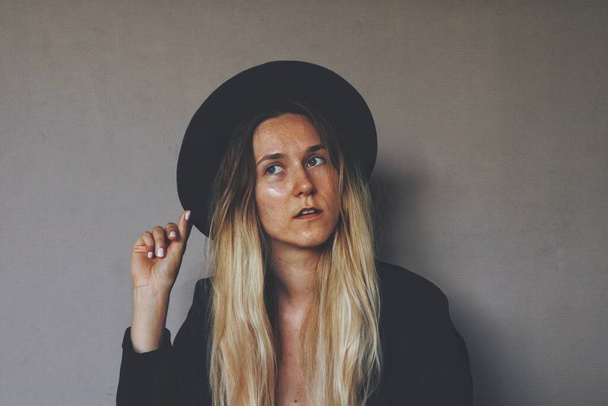 Young blond female wiccan witch wearing a black witch hat and black blazer. Artsy portrait against dark grey wall. Woman holding a hat brim with her right hand fingers, looking away from camera lens - Photo, Image